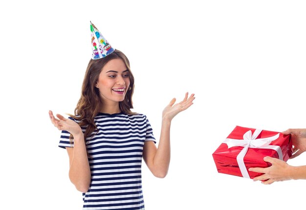Całkiem młoda kobieta w koszulce w paski i czapce z okazji uroczystości, trzymająca czerwony prezent i kolorowe balony