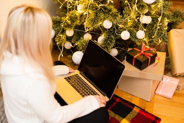 Zdjęcie całkiem młoda kobieta w modnej zimowej mody, siedząc na podłodze ze skrzyżowanymi nogami, trzymając gadżet laptopa przed ozdobą choinkową, patrząc w kamerę.