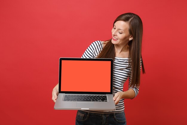Całkiem młoda kobieta w paski ubrania patrząc na komputerze typu laptop z pustym czarnym pustym ekranem w rękach na białym tle na czerwonym tle. Ludzie szczere emocje, koncepcja stylu życia. Makieta miejsca na kopię.