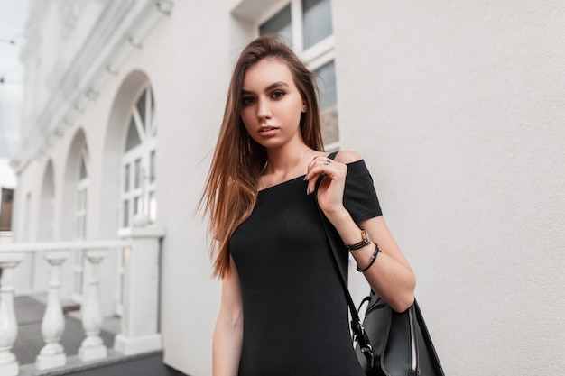 Całkiem seksowna młoda modelka w eleganckiej czarnej sukience ze skórzanym stylowym czarnym plecakiem podróżuje po mieście. Atrakcyjna modna dziewczyna lubi spacer po ulicy. Uliczny styl młodzieżowy.