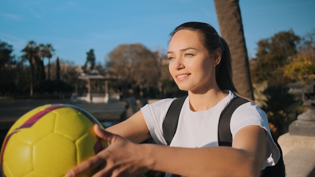 Całkiem sportowa dziewczyna z plecakiem i piłką nożną szczęśliwie odpoczywa w pięknym parku