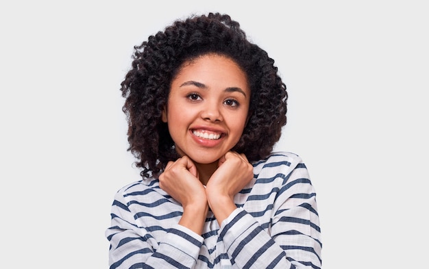 Całkiem szczęśliwa African American młoda kobieta z pozytywnym uśmiechem trzymająca ręce na brodzie patrząc w kamerę ma radosny wyraz Urocza ciemnoskóra kobieta ma pozytywne emocje
