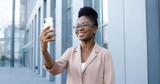 Całkiem Szczęśliwy African American Młoda Kobieta W Okularach O Wideoczat Na Smartfonie Na świeżym Powietrzu W Budynku Biznesowym. Radosna Piękna Kobieta Rozmawia I Prowadzi Wideoczat Przez Kamerę Internetową W Telefonie Komórkowym.