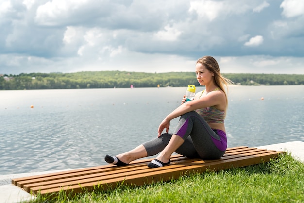 Zdjęcie całkiem szczupła kobieta ubrana w sportowe ubrania, siedząca nad jeziorem i relaksująca po wysiłku po ciężkim dniu pracy
