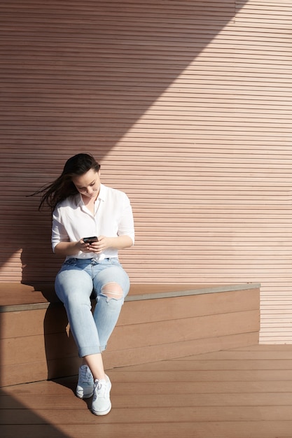 Całkiem uśmiechnięta młoda kobieta siedzi na zewnątrz w wietrzny dzień, wysyłając SMS-y do znajomych i sprawdzając posty w mediach społecznościowych