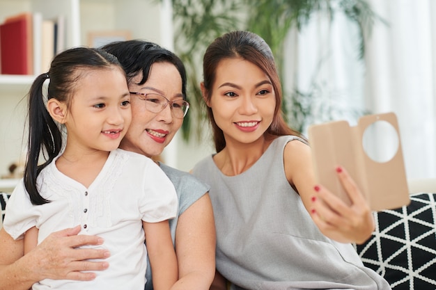 Całkiem uśmiechnięta młoda kobieta ze smartfonem robi selfie ze swoją starszą matką i małą córeczką