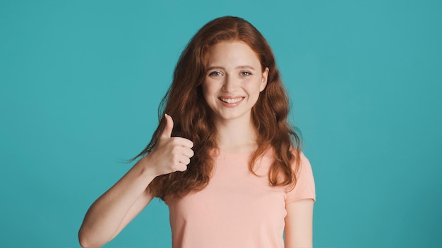 Całkiem wesoła rudowłosa dziewczyna szczęśliwie pokazująca kciuk w górę gestem przed kamerą na kolorowym tle Dobrze zrobione wyrażenie