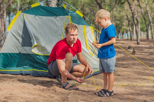 Camping ludzie outdoor lifestyle turyści rozbijają swój zielony szary kemping w letnim lesie w pobliżu lazurowego morza Słodki chłopiec syn pomaga ojcu składać namiot Naturalna edukacja dzieci