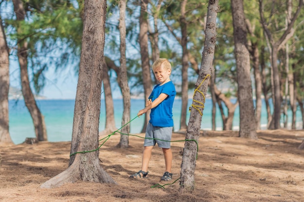 Camping ludzie outdoor styl życia turyści w letnim lesie w pobliżu lazurowego morza Blond poważny chłopiec w niebieskiej koszulce nauka technik przetrwania praktyka metody wiązania węzłów naturalna edukacja dzieci