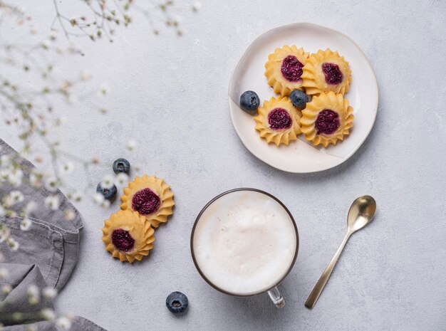 Cappuccino lub latte z pianką mleczną w filiżance z ręcznie robionymi ciasteczkami jagodowymi i jagodami na jasnym tle z gałązkami łyszczec Koncepcja wiosenne śniadanie rano