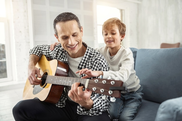 Cenny czas. Przystojny radosny ciemnowłosy uśmiechnięty i grający na gitarze mężczyzna oraz jego syn siedzący za nim na kanapie