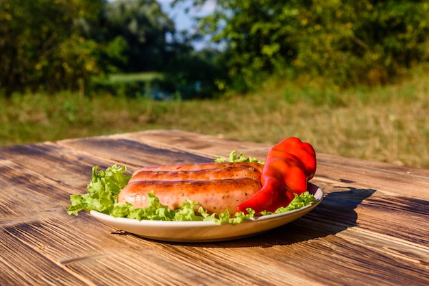 Ceramiczny talerz z grillowanymi kiełbaskami, liśćmi papryki i sałaty na rustykalnym drewnianym stole