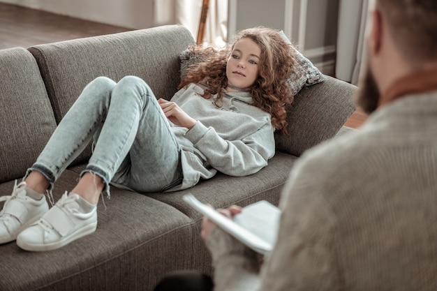 Chłodzenie na kanapie. Kręcona nastolatka w szarej bluzie z kapturem odpoczywa na kanapie i rozmawia z doradcą