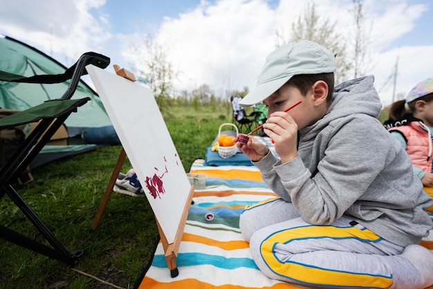 Chłopiec cieszący się świeżym powietrzem na kocu piknikowym i malujący na płótnie w ogrodowym wiosennym relaksie w parku