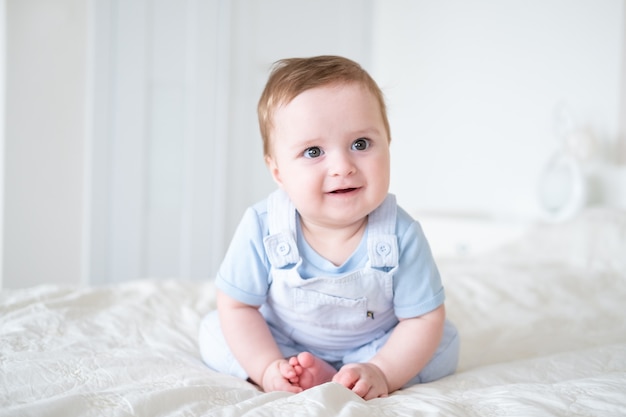 Chłopiec dziecko 6 miesięcy w ubraniach blu, uśmiechając się i siedząc na białym łóżku w domu.