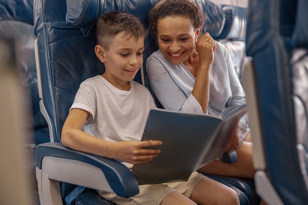 Chłopiec, dziecko na pokładzie trzyma książkę i przegląda ją razem z mamą podczas lotu. Załoga pokładowa obsługująca rodzinę w samolocie. Koncepcja transportu lotniczego i turystyki