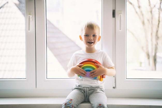Chłopiec dziecko na tle okna trzyma drewnianą tęczową zabawkę
