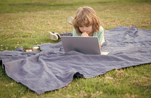 Chłopiec dziecko surfuje po Internecie w laptopie relaksuje się na kocu leżącym na trawie naturalnej na zewnątrz edukacja na odległość