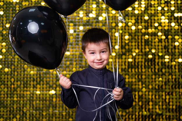 Chłopiec dziecko trzyma czarne balony na tle ze złotymi błyszczącymi cekinami, paillettes.