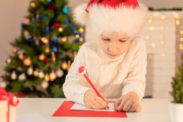 Chłopiec dziecko w czerwonym kapeluszu Świętego Mikołaja pisze list do Świętego Mikołaja Boże Narodzenie lub nowy rok przytulne wakacje koncepcja Boże Narodzenie czas Selektywna ostrość