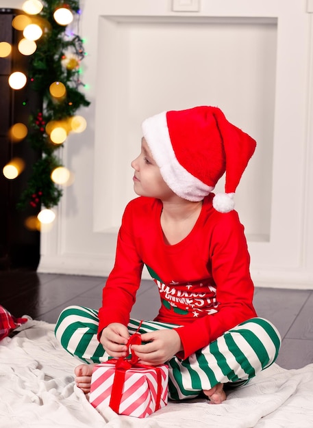 Chłopiec dziecko w zabawnej czapce Mikołaja i świątecznej piżamie siedzi na kocu z prezentem w dłoniach