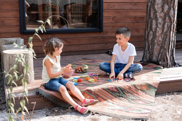 Chłopiec I Dziewczynka Bawią Się Na Ganku W Pobliżu Drewnianego Domu, Siedzą Na Haftowanym Dywanie, Komunikują Się I Grają W Gry Edukacyjne