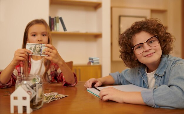 Chłopiec i dziewczynka siedzą przy stole i wkładają oszczędności do słoika