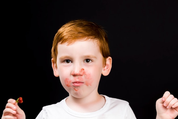 Chłopiec je dojrzałe truskawki i posmarowany czerwonymi jagodami, zbliżenie portret dziecka podczas jedzenia deseru z jagód