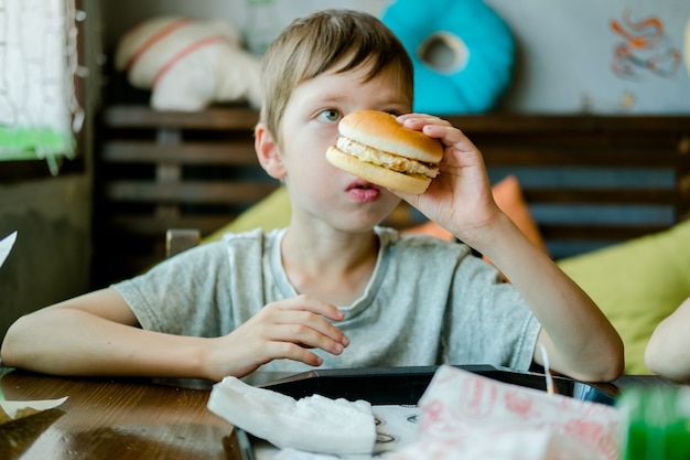 Chłopiec je duży burger z kotletem. Hamburger w rękach dziecka. Pyszny i satysfakcjonujący burger z kotleta z kurczaka. Jedzenie na wynos