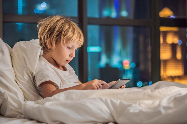 Chłopiec korzysta z tabletu w swoim łóżku przed pójściem spać na tle nocnego miasta Dzieci i koncepcje technologiczne