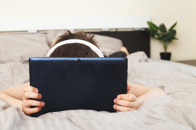 Chłopiec leżący na łóżku w domu zakrył twarz tabletem PC i czytał online