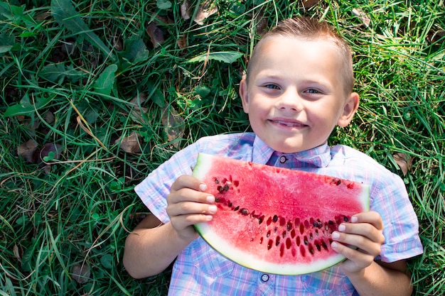Chłopiec leży na zielonej trawie i trzyma duży kawałek arbuza, mały uczeń je arbuza w słoneczny letni dzień, miejsce