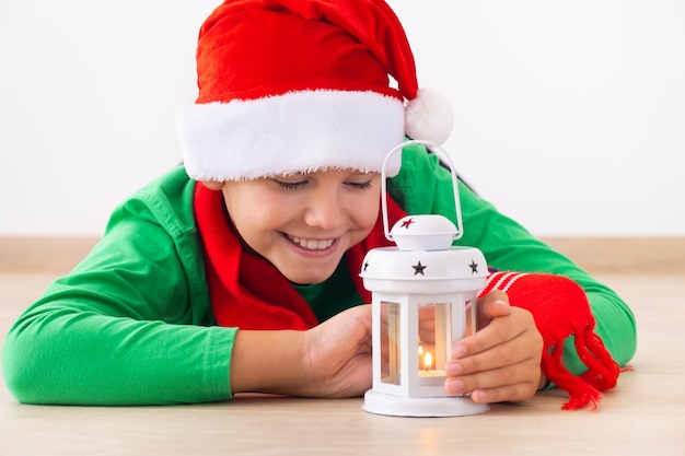 Chłopiec ma na sobie czapkę Mikołaja i zimowy szalik Dziecko leży na podłodze i patrzy na latarnię bożonarodzeniową i ogrzewa ręce