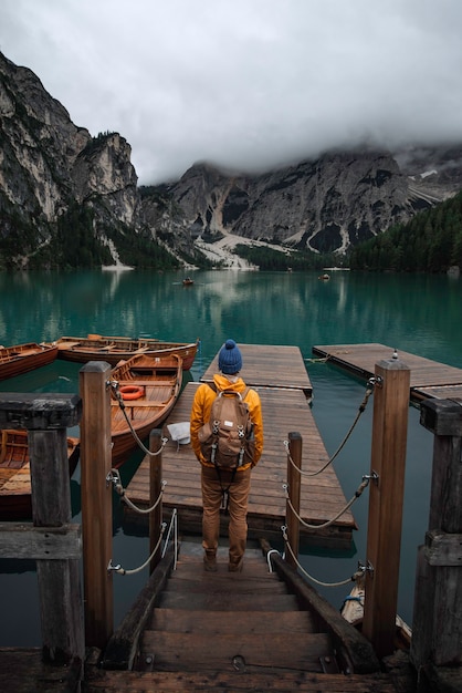 Chłopiec młodego podróżnika z niebieskim kapeluszem, plecakiem vintage i żółtym płaszczem przeciwdeszczowym na molo w lago di Braies otoczonym górami włoskich Alp