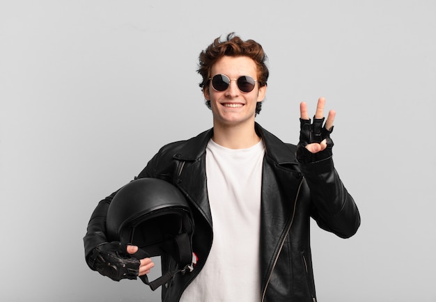 Chłopiec motocyklista uśmiechający się i patrzący przyjaźnie, pokazujący numer trzy lub trzeci z ręką do przodu, odliczający w dół