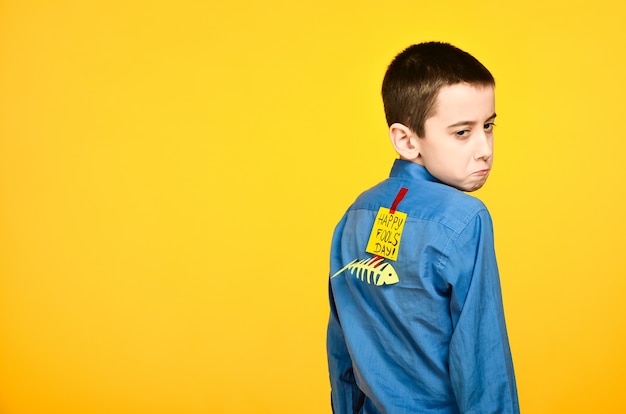 Chłopiec na żółtym tle w niebieskiej koszuli z przyklejoną taśmą rybną i kartką papieru na plecach