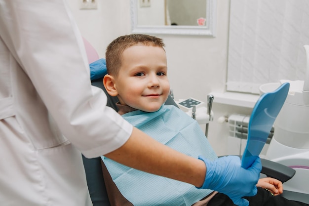 Chłopiec Patrzy W Lustro Z Uśmiechem Zębów, Siedząc Na Krześle Z Dentystą W Gabinecie Stomatologicznym.