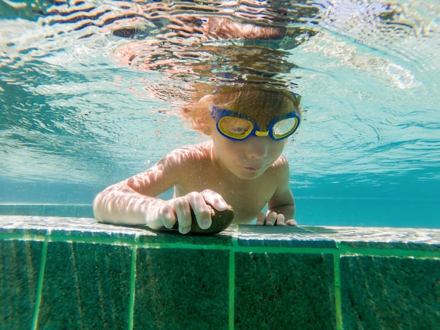 Chłopiec pływa pod wodą w basenie, uśmiechając się i wstrzymując oddech, w okularach do pływania