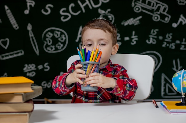 Chłopiec przedszkolak odrabiania lekcji w szkole. Szkolny chłopiec z szczęśliwym wyrazem twarzy w pobliżu biurka z przyborami szkolnymi. Edukacja. Nauka przede wszystkim. Koncepcja szkoły.