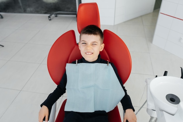 Chłopiec siedzi na czerwonym fotelu dentystycznym i uśmiecha się w nowoczesnej białej stomatologii Leczenie i profilaktyka próchnicy od dzieciństwa Nowoczesna stomatologia i protetyka