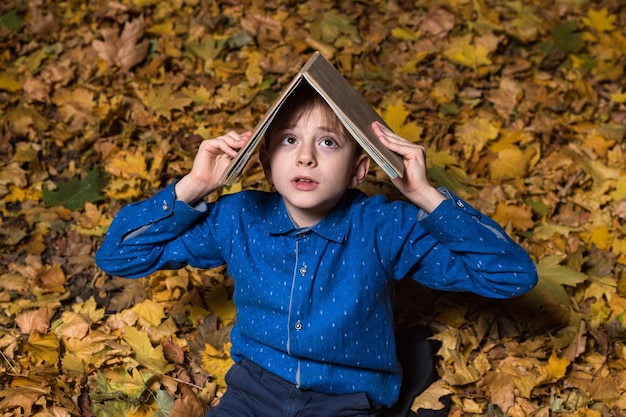 Chłopiec siedzi na opadłych jesiennych liściach i kładzie książkę na głowie horror book