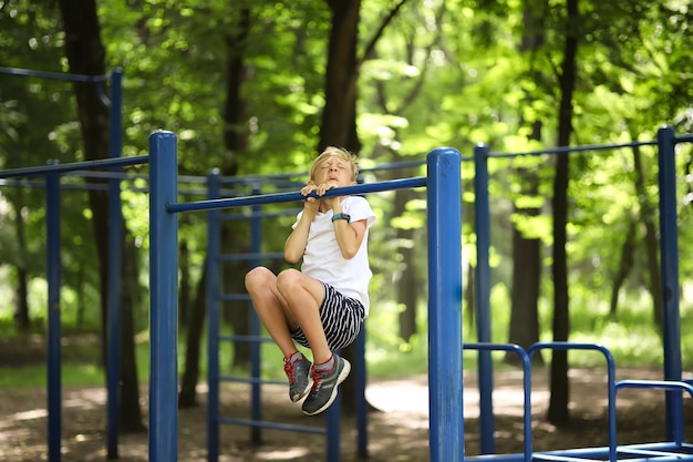 Chłopiec sportowiec w parku podciąga się na drążku, skrzywił się i podciąga z wysiłkiem