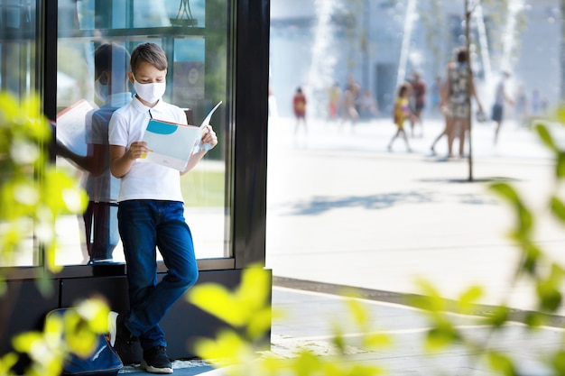 Zdjęcie chłopiec-student w masce czyta książkę na placu miejskim podczas pandemii covid 19