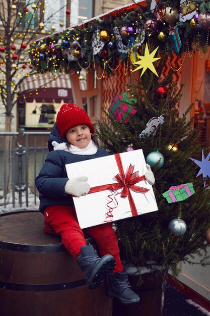 Zdjęcie chłopiec świąteczny dziecko w ciepłych ubraniach trzyma prezent i siedzi na beczce obok choinki