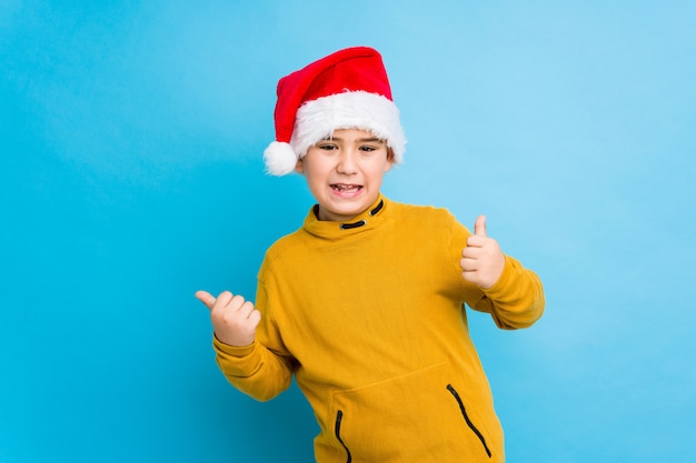 Chłopiec świętuje boże narodzenie dzień jest ubranym Santa kapelusz odizolowywającego podnoszący oba aprobaty, uśmiechnięty i ufny.