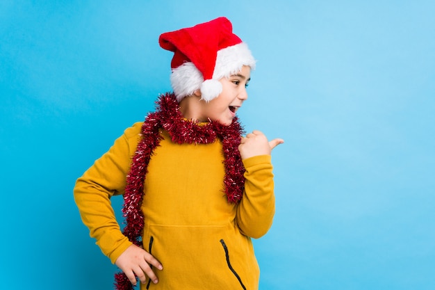 Chłopiec świętuje boże narodzenie dzień jest ubranym Santa kapelusz odizolowywających punkty z kciuka palcem daleko od, śmia się i beztroski.