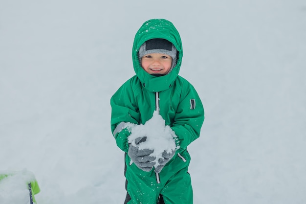 Chłopiec trzyma w rękach grudkę śniegu i uśmiecha się chłopiec ubrany jest w ciepły zielony kombinezon