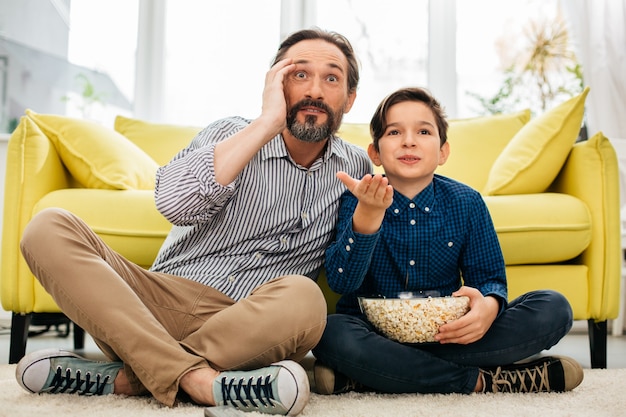 Chłopiec Trzymający Popcorn I Jego Ojciec Wyglądający Na Zachwyconego Siedząc Na Podłodze I Oglądając Niesamowity Mecz W Telewizji