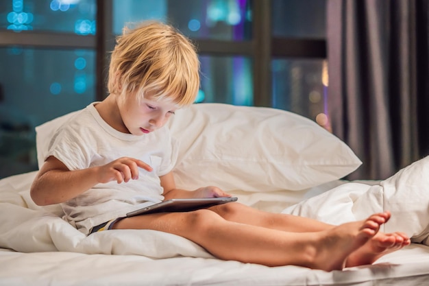 Chłopiec Używa Tabletu W Swoim łóżku Przed Pójściem Spać Na Tle Nocnego Miasta. Koncepcje Dotyczące Dzieci I Technologii