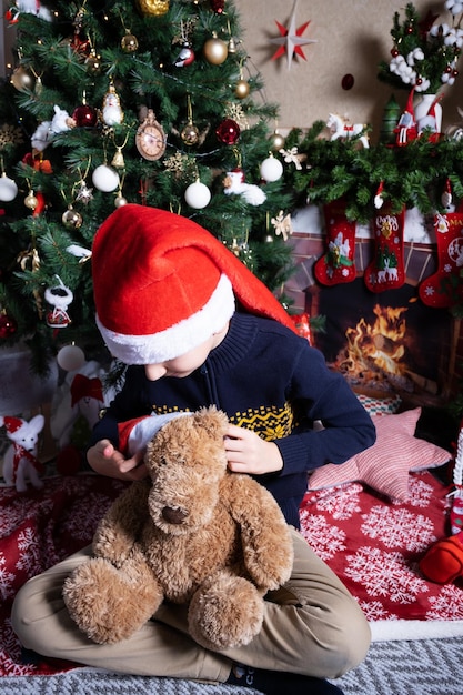 Chłopiec w czapce Świętego Mikołaja siedzi na podłodze przy kominku zakłada świąteczną czapkę na zabawkę-miś